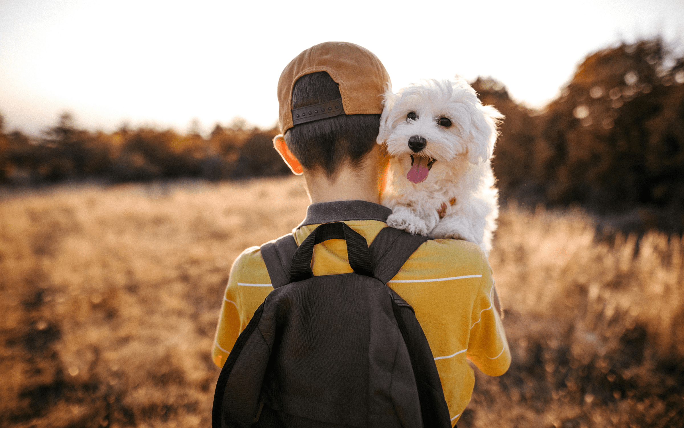 A child in a heat and a yellow shirt with a backpack, carrying a small white dog